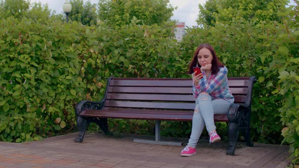 Young Woman with Mobile Phone Rests Sitting on Park Bench Surrounded By Bushes