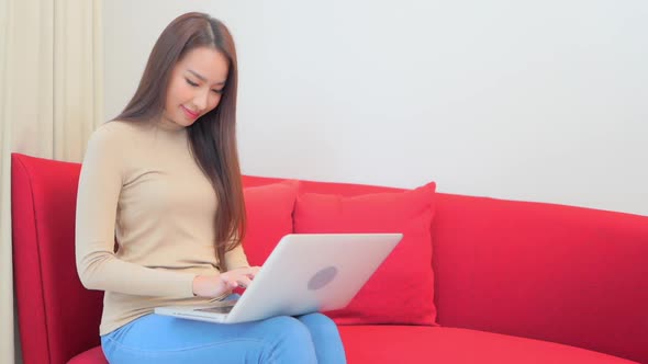 Asian woman in 30th typing on laptop computer holding it on laps sitting on red home sofa