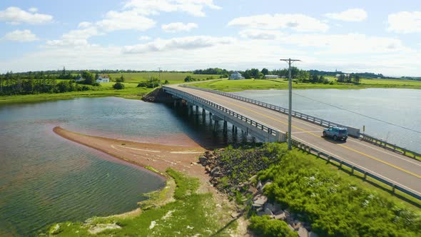 Aerial view follows vehicle over bridge in Prince Edward Island on a summer day.