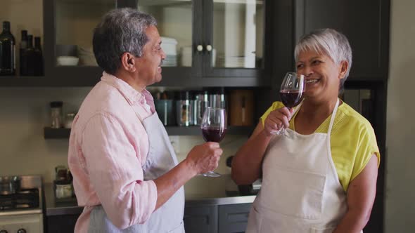 Happy senior mixed race couple wearing aprons toasting with wine in kitchen