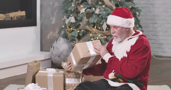 Old Caucasian Man with White Beard and Mustaches Holding Gift Box and Shaking It. Santa in Red Hat