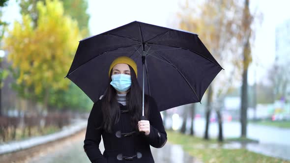 A Young Woman in a Protective Mask Walking in the Park Under Umbrella. Rainy Day, During Second Wave