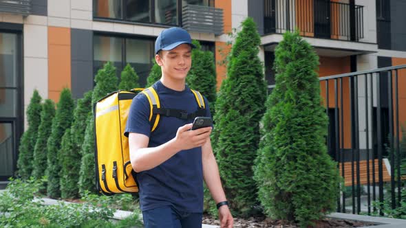 Smiling Food Delivery Man Courier Walks Street Watching Navigation in Smartphone