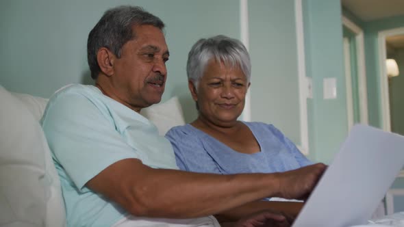 Happy senior mixed race couple sitting in bed using laptop