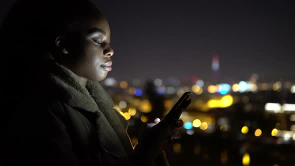 A Young Black Woman Works on a Smartphone in an Urban Area at Night  Side Closeup