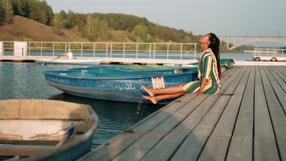 Beautiful AfricanAmerican Girl with Dreadlocks in a Bright Dress Sits on a Pier Among the Boats with