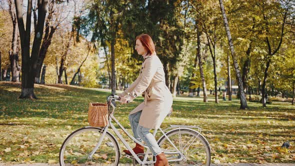 Young Woman is Riding Bicycle with Basket By Tiled Path of Autumn City Park
