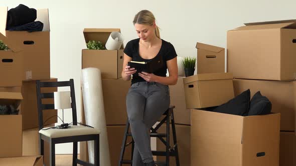 A Moving Woman Sits on a Chair in an Empty Apartment and Writes Into a Notebook, Surrounded By Boxes