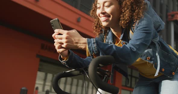 Happy biracial woman in city, sitting on bike using smartphone