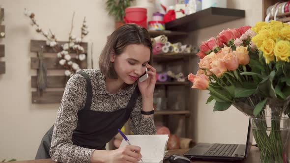 Cheerful Saleswoman of Flowers Shop Is Talking By Phone and Working on Laptop