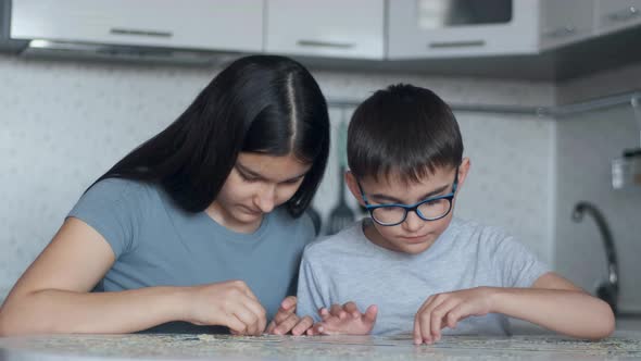 Cheerful Brother and Sister Assemble a Puzzle While Sitting at a Table at Home in the Kitchen. Close