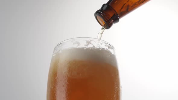 Beer is pouring with bubbles and foam in large beer glass from bottle on white background close up.