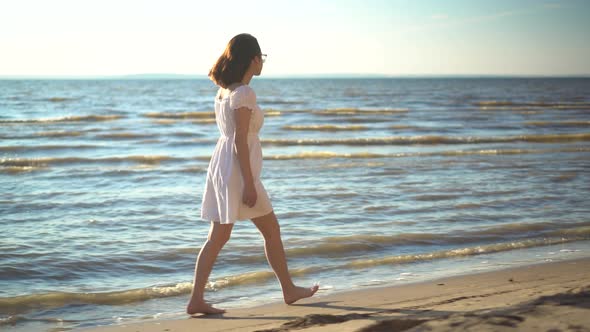 Attractive Young Woman Walks Along the Sea Beach. A Girl in a White Dress Walks Barefoot Along the