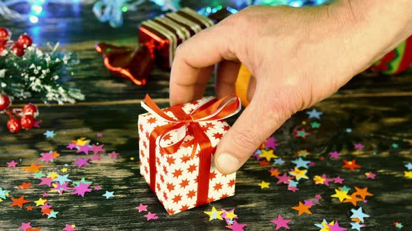 Hand Puts Gift Box of Red Bow on Table Decorated with Christmas Stuff on Bokeh Garland