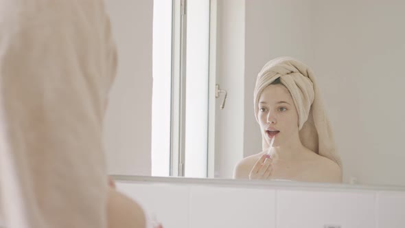 Teenage girl applying lip balm and lip gloss in front of the bathroom mirror
