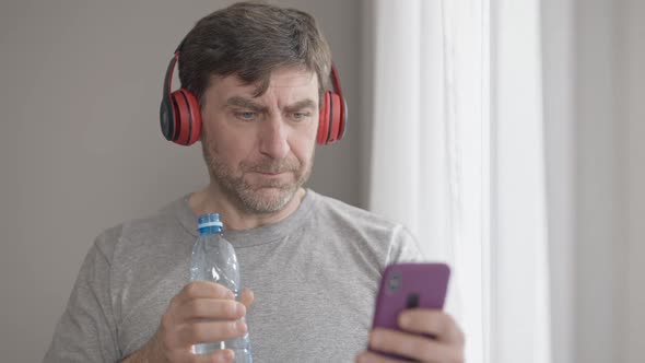 Positive Middle Aged Man in Headphones Drinking Refreshing Water and Listening to Playlist 