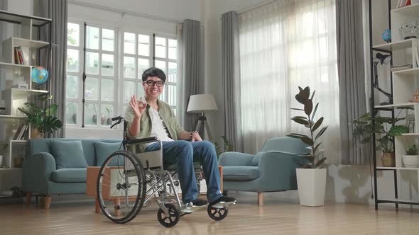 Asian Man Sitting In A Wheelchair Smiling And Showing Ok Sign In Living Room