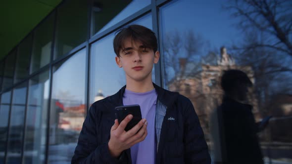 Medium Shot of Handsome Young Man Working on Smartphone Stands on the Street in Front of a Modern