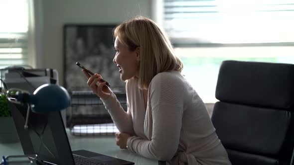 A Close-up Profile of a Middle-aged Woman with Blond Hair, Sitting in an Office at a Laptop, Smiling