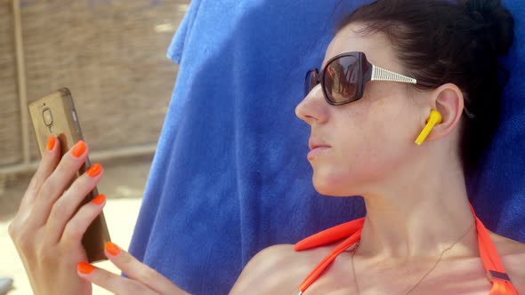 Portrait, a Young Woman in Sunglasses and Bluetooth Earphones Is Watching Video From Smartphone