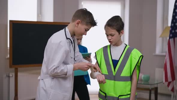 Caucasian Teenage Boy in Doctor Gown Bandaging Hand of Friend in Builder Uniform