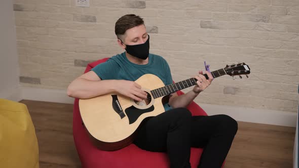 Young Man with a Black Medicinal Mask on Face Is Playing at the Guitar While Is Sitting Comfortably