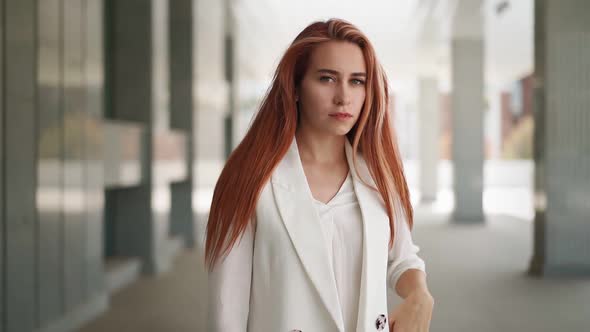 Beautiful modern business woman with long red hair in trendy white coat stands near office building