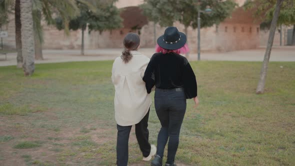 Rear Tracking View of Two Stylish Girls Wandering Around a Park Holding Hands