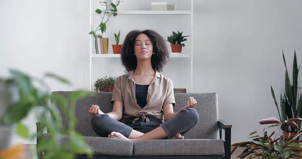 Calm Relaxed African Woman Sitting at Home on Sofa with Closed Eyes, Crossed Legs, Meditating, Doing