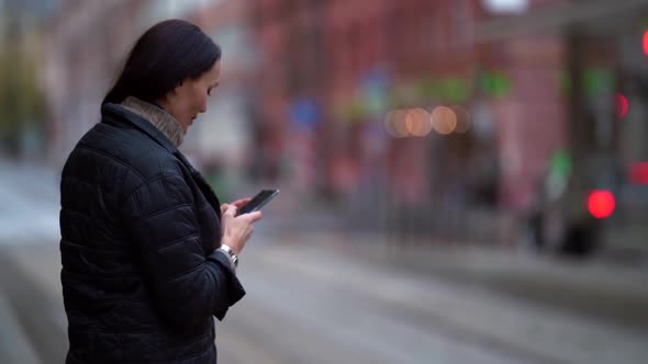 a Brunette in a Knitted Sweater and a Dark Coat Stands on a City Street and Types a Message on Phone