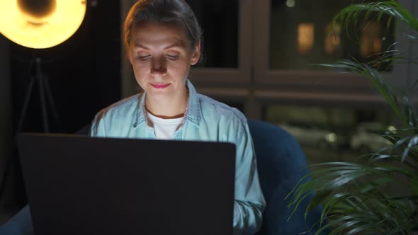 Woman is Sitting in the Armchair and Working on a Laptop at Night
