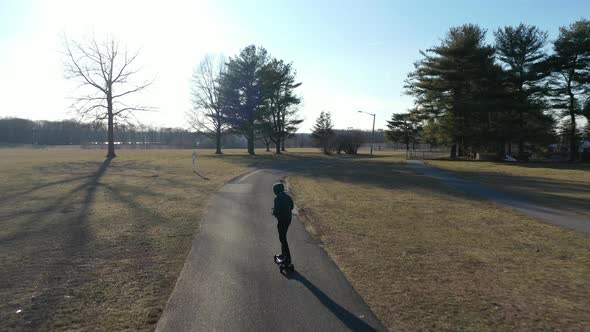 An aerial tracking of a man on an electric skateboard in an empty park on a sunny day. The drone fol