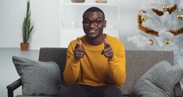 Portrait of Afro American Guy in Glasses and Yellow Sweater Sits on Couch at Home Near Christmas