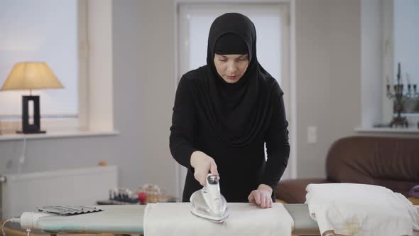 Portrait of Beautiful Muslim Lady in Traditional Clothes Ironing and Folding Laundry at Home