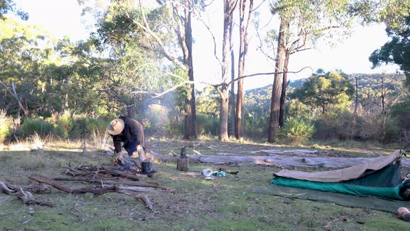 A bushman puts a billy on the boil with his swag set up in the Australian bush.