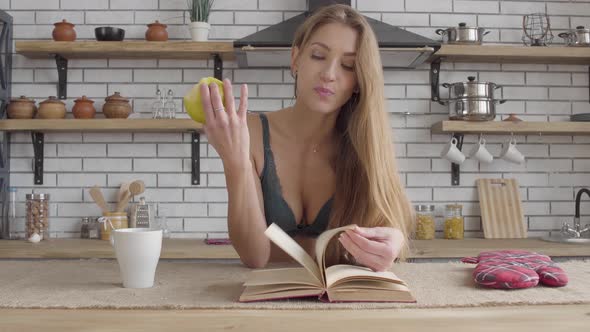 Portrait Young Sensual Woman in Lingerie Reading Book and Eating Green Apple in the Kitchen. Healthy