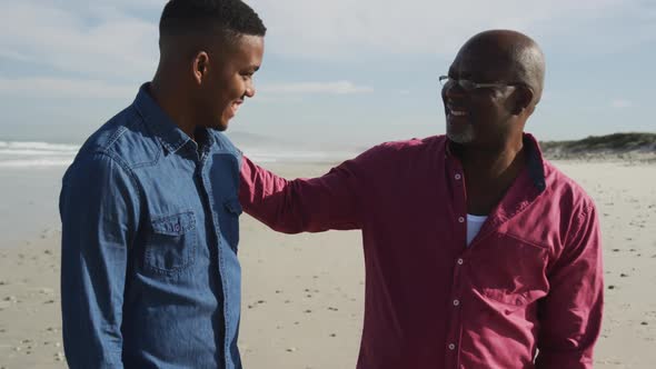 African american father standing on beach with teenage son, putting hand on his shoulder and smiling