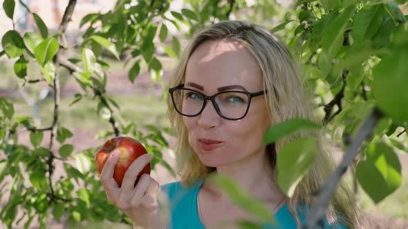 portrait of a young woman biting and eating a ripe red apple, looking at the camera in the garden