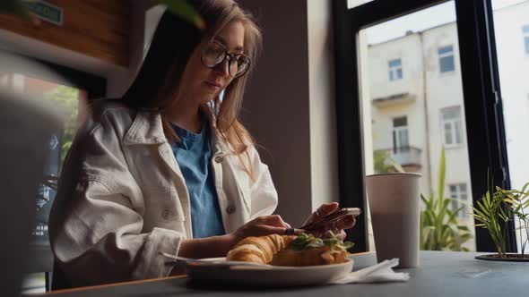 Front View of Attractive Woman Wearing Glasses Using Smartphone, Drinking Coffee in Cafe