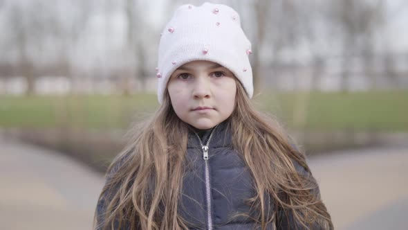 Close-up Face of Cute Caucasian Girl in Coat and Hat Looking at Camera. Portrait of Serious Brunette