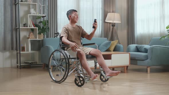 Young Asian Boy Sitting In A Wheelchair While Waving Hand Video Calling Distance In Phone At Home