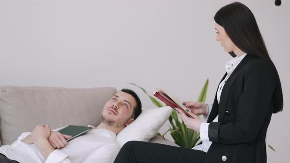 The Man Is Laying on the Bed with a Book and Talking To a Woman Sitting Next To Him