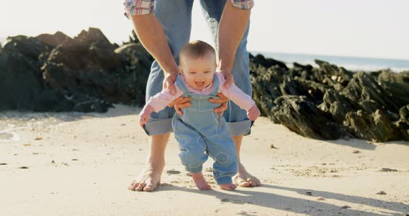 Front view of mid adult caucasian father helping baby to walk at beach on a sunny day 4k