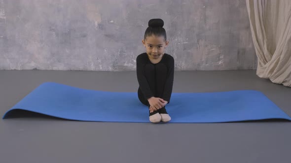 Little Asian Gymnastic Girl in Black Bodysuit Sitting on Yoga Mat and Looking on Camera