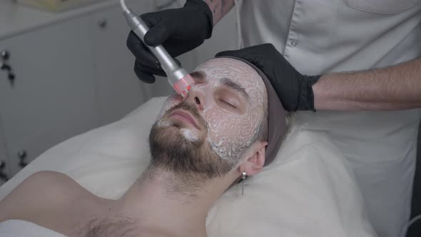 Face of Caucasian Man with Closed Eyes with Beautician Cleansing Nose Skin with Red LED Light Device