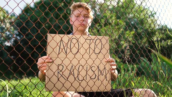 a Young Caucasian Teenager Man in a Blue Shirt Sits Behind a Metal Lattice Fence with a Corton Box