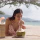 Young Woman Sitting at Table with Coconut on Beach - VideoHive Item for Sale