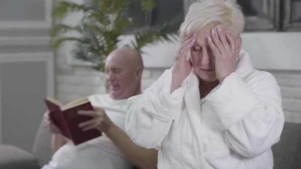 Portrait of Sad Caucasian Woman with Headache Sitting in White Bathrobe and Rubbing Temples