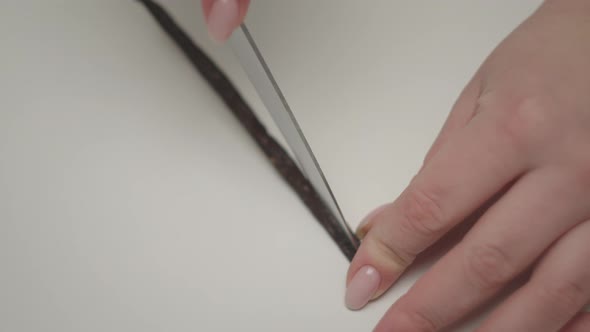 Female hands cutting vanilla pod with knife on white table in slow motion.