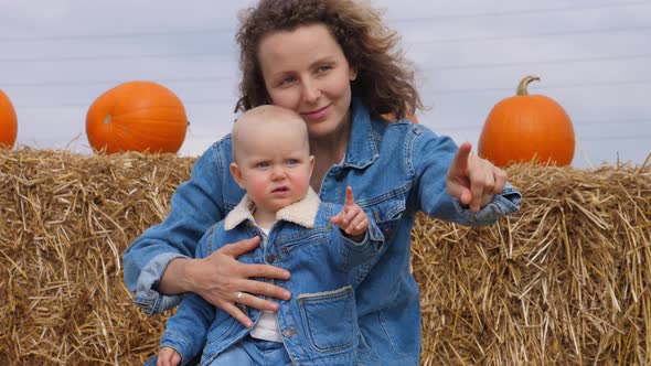 Portrait of Caucasian Mom and Baby in Matching Denim Looks Sitting on a Hay Bale Pointing at